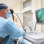 A gastroenterologist holding an endoscope beside a monitor in a procedure room