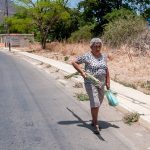 An older woman walking along a sunny roadside carrying a shopping bag