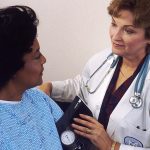 Doctor with a stethoscope and blood pressure cuff speaking with a patient in a hospital gown.