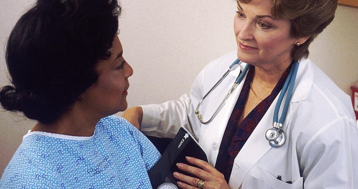 Doctor with a stethoscope and blood pressure cuff speaking with a patient in a hospital gown.